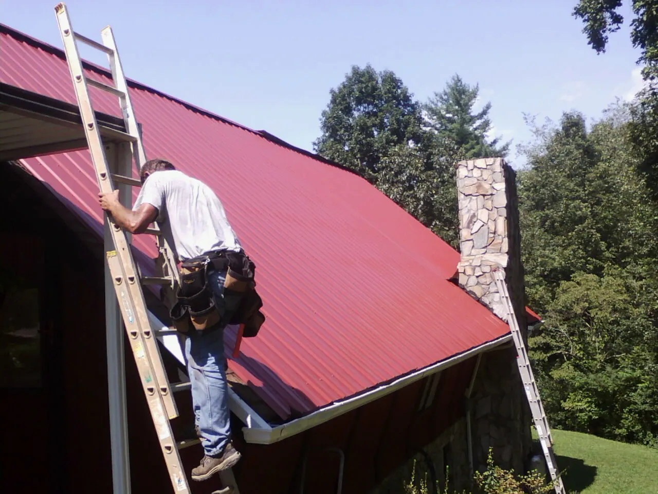Roofer on a ladder beside a red metal roof and stone chimney — Soffit & Fascia Repair by Roofing Brothers in Winthrop Harbor